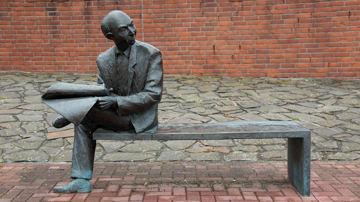 Statue: Mann sitzt auf Bank und hält eine Zeitung in der Hand