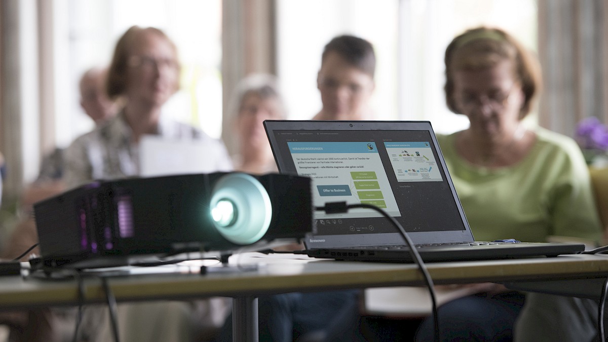 Blick auf einen Beamer und ein Notebook. Im Hintergrund sitzen die Teilnehmenden des Workshops.